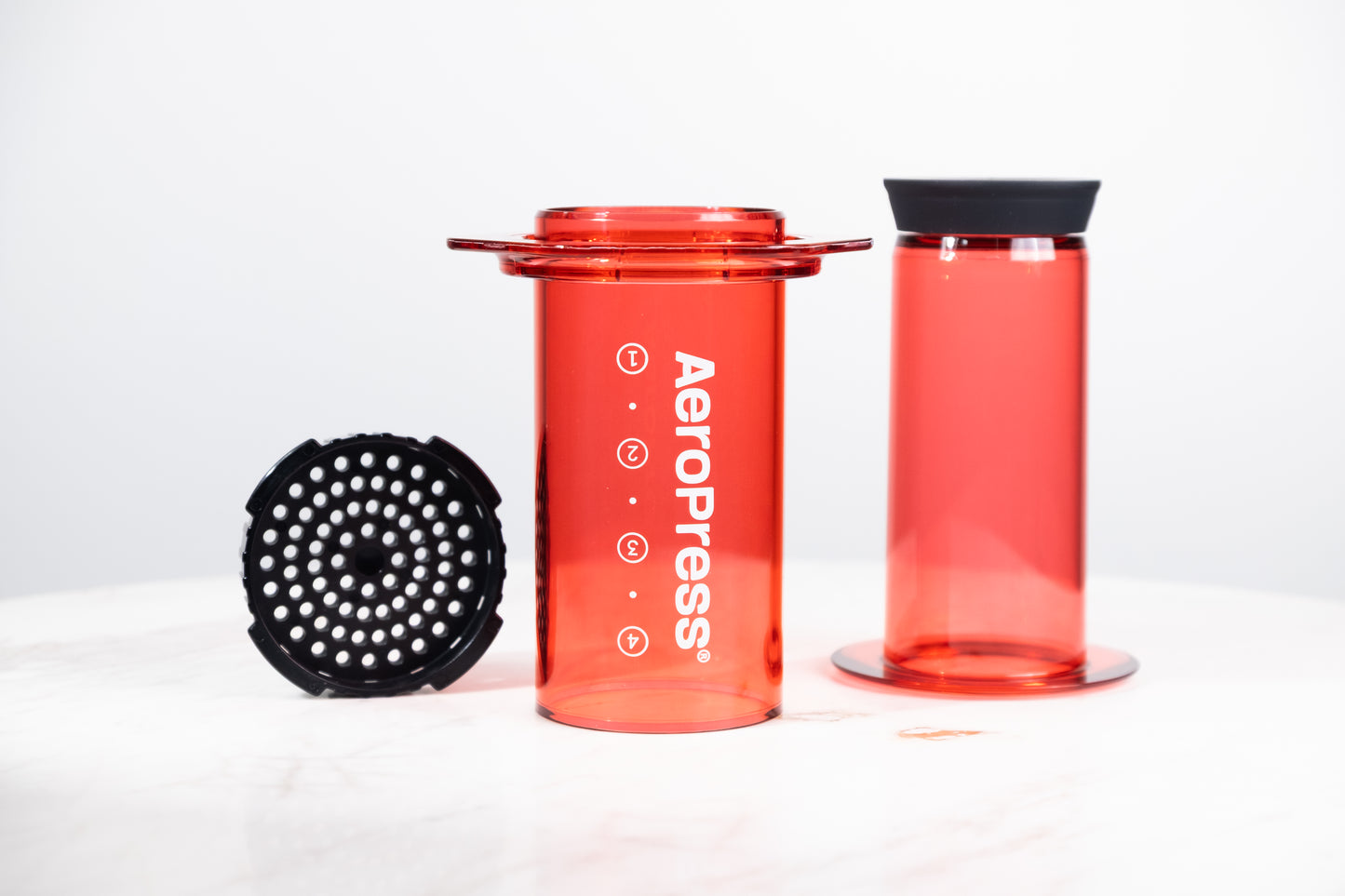 Cafetera manual AeroPress Clear en color rojo translúcido. Despiece del método de preparación que incluye cámara, émbolo y tapa de filtro para café de especialidad.