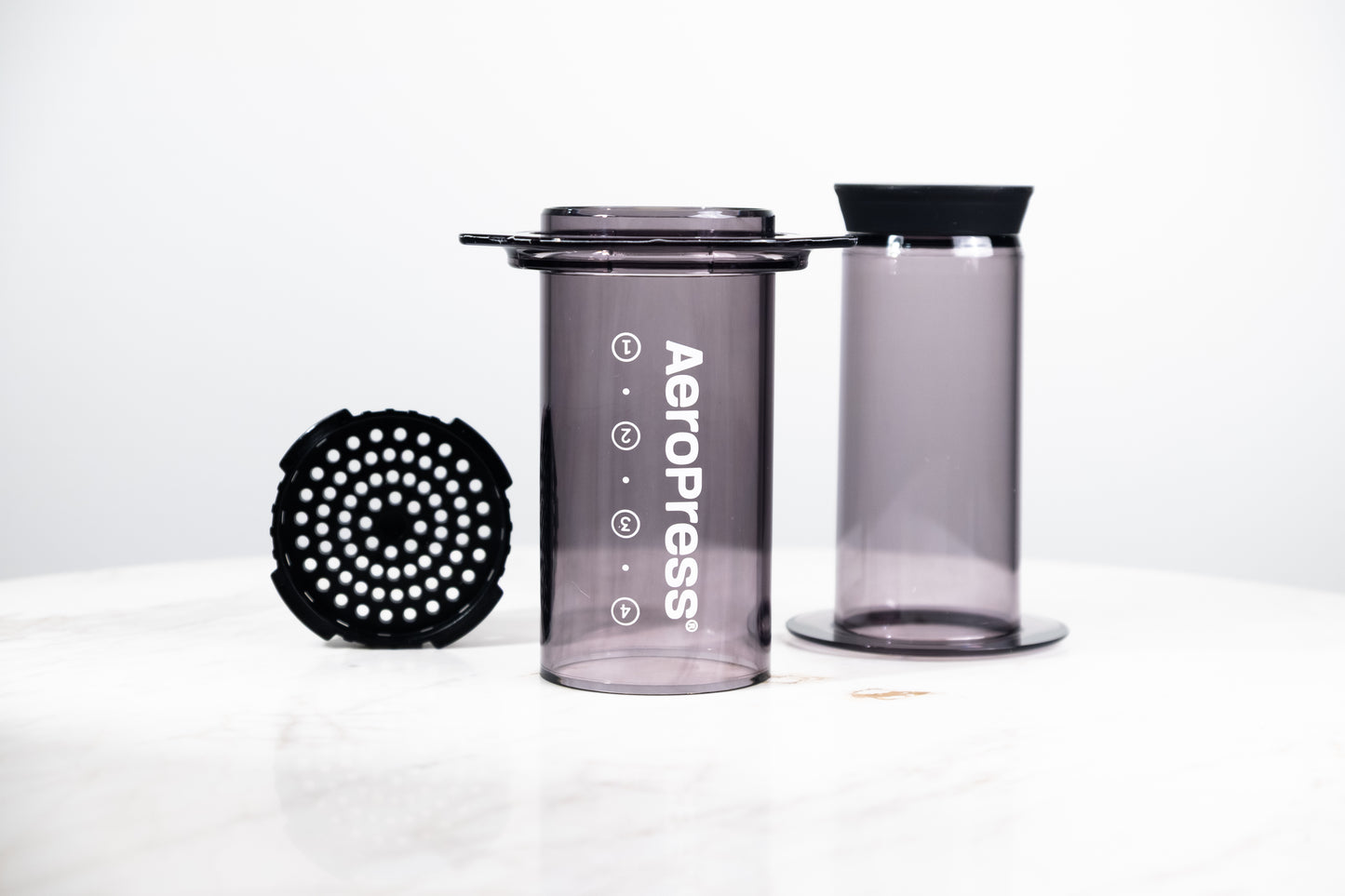 Cafetera manual AeroPress Clear en color negro translúcido (Smoke). Despiece del método de preparación que incluye cámara, émbolo y tapa de filtro para café de especialidad.