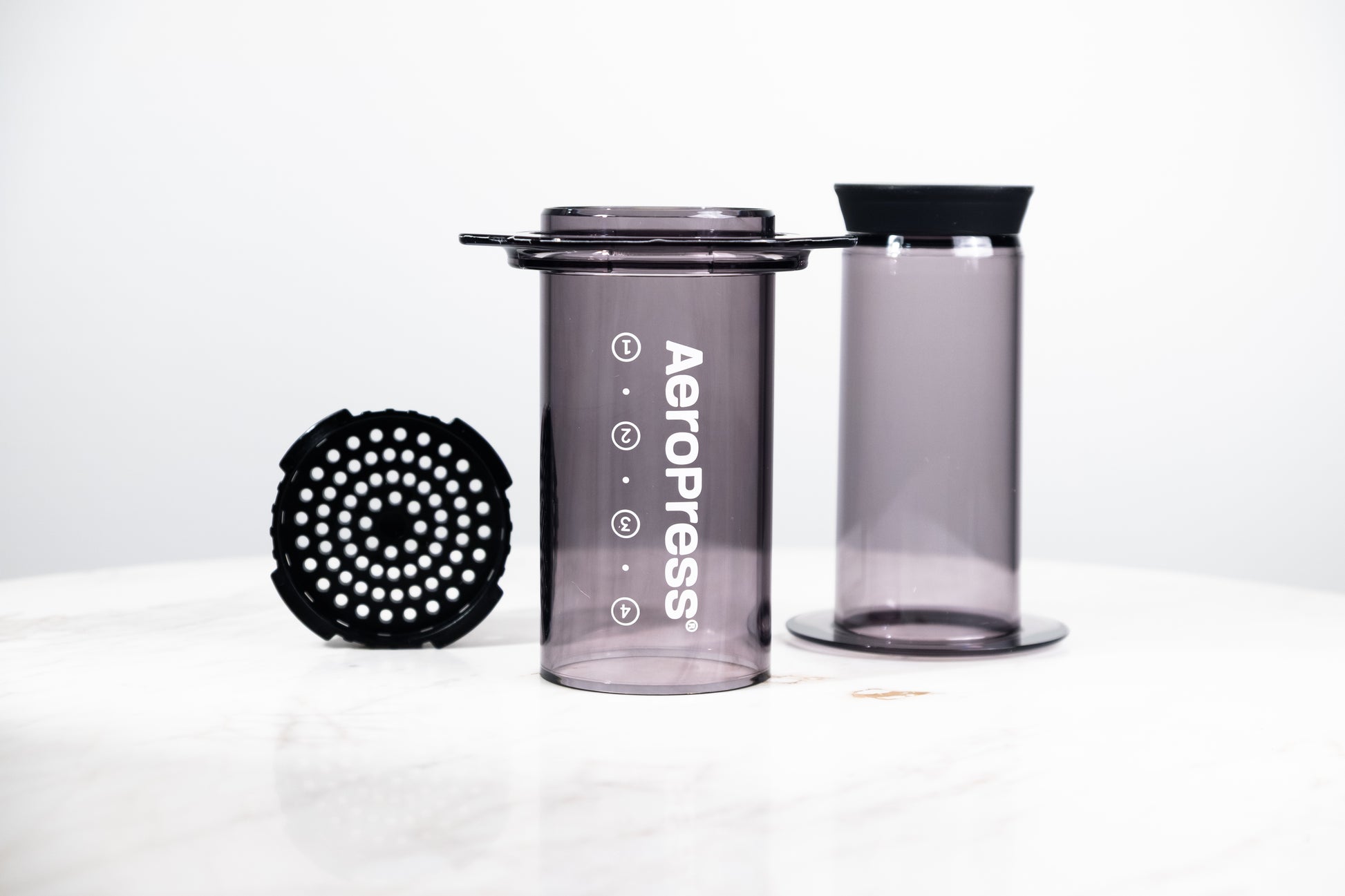 Cafetera manual AeroPress Clear en color negro translúcido (Smoke). Despiece del método de preparación que incluye cámara, émbolo y tapa de filtro para café de especialidad.