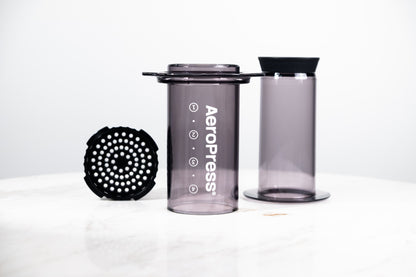 Cafetera manual AeroPress Clear en color negro translúcido (Smoke). Despiece del método de preparación que incluye cámara, émbolo y tapa de filtro para café de especialidad.
