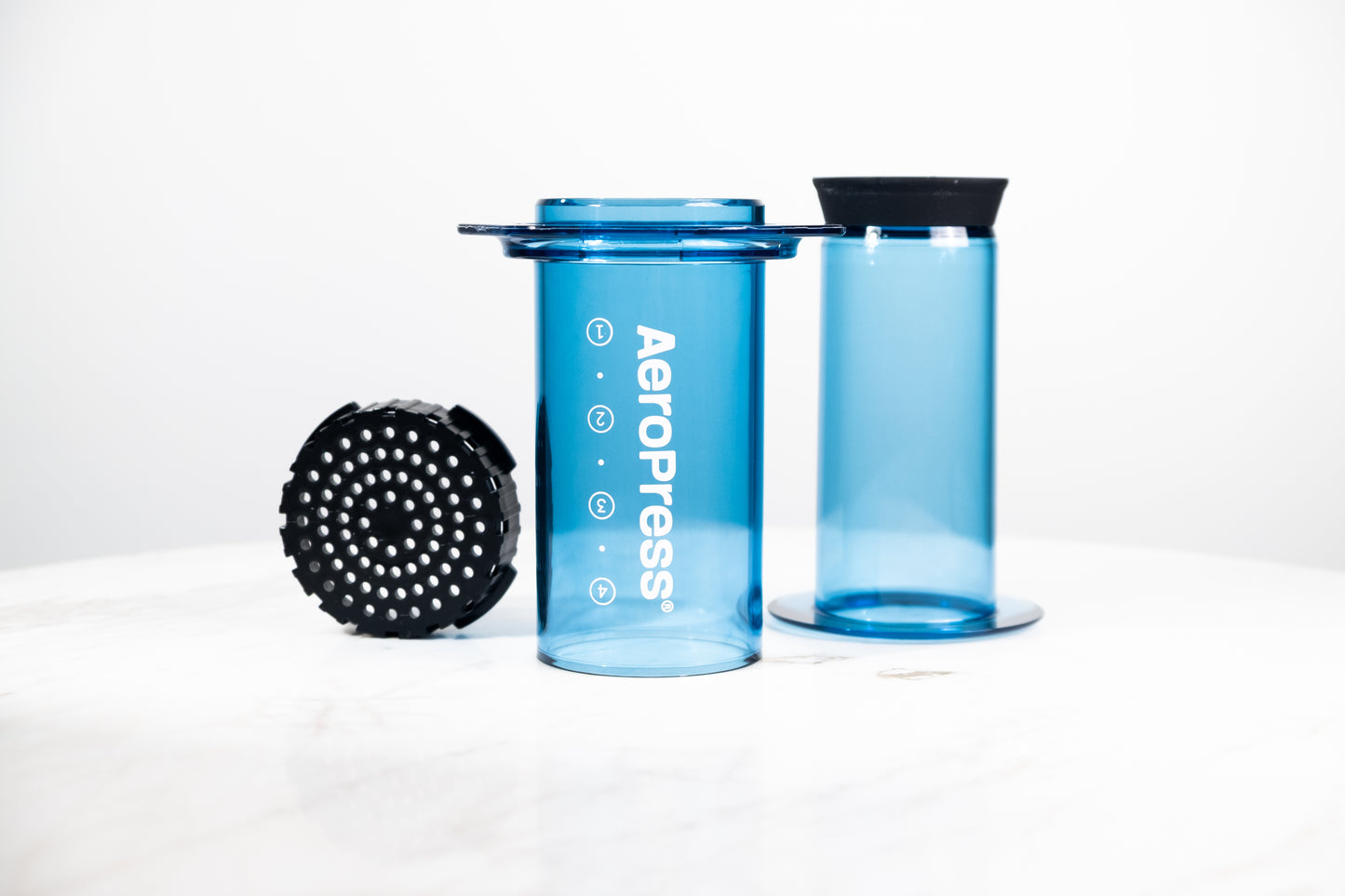 Cafetera manual AeroPress Clear en color azul translúcido. Despiece del método de preparación que incluye cámara, émbolo y tapa de filtro para café de especialidad.