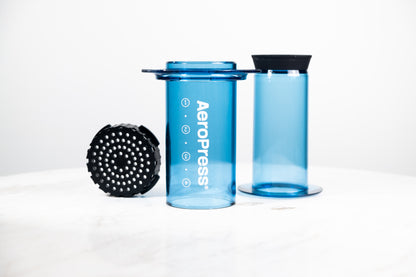 Cafetera manual AeroPress Clear en color azul translúcido. Despiece del método de preparación que incluye cámara, émbolo y tapa de filtro para café de especialidad.