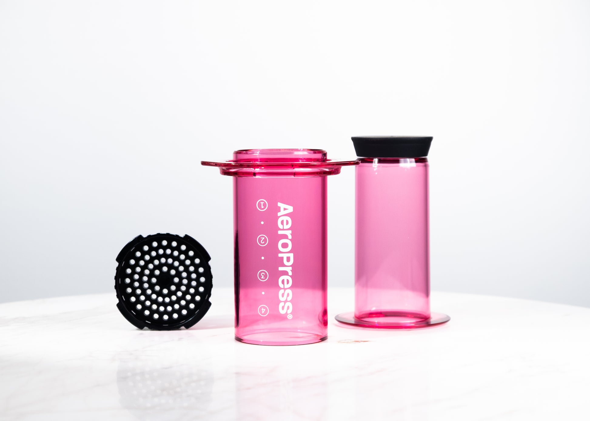 Cafetera manual AeroPress Clear en color fucsia translúcido. Despiece del método de preparación que incluye cámara, émbolo y tapa de filtro para café de especialidad.
