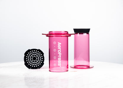 Cafetera manual AeroPress Clear en color fucsia translúcido. Despiece del método de preparación que incluye cámara, émbolo y tapa de filtro para café de especialidad.