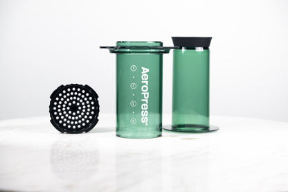 Cafetera manual AeroPress Clear en color verde translúcido. Despiece del método de preparación que incluye cámara, émbolo y tapa de filtro para café de especialidad.