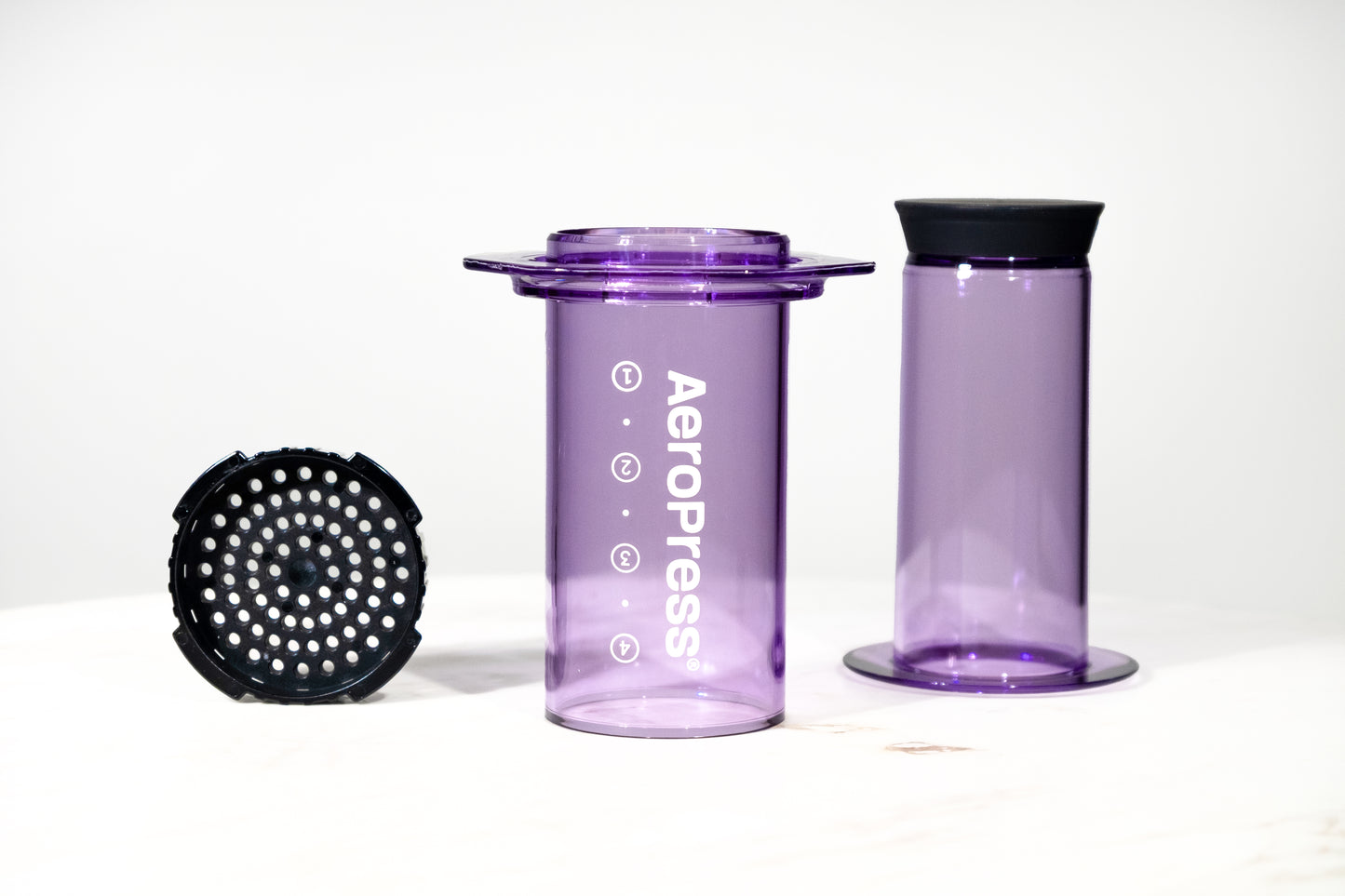 Cafetera manual AeroPress Clear en color morado translúcido. Imagen detallada de las piezas desmontadas: cámara, émbolo y tapa de filtro para preparar café de especialidad.