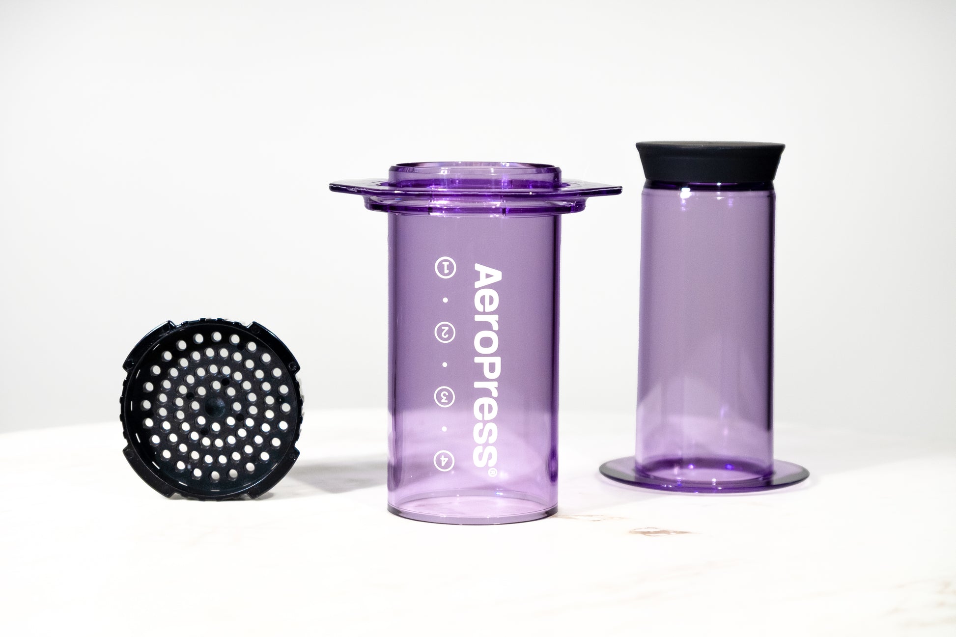 Cafetera manual AeroPress Clear en color morado translúcido. Imagen detallada de las piezas desmontadas: cámara, émbolo y tapa de filtro para preparar café de especialidad.