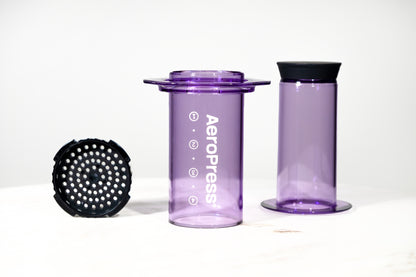 Cafetera manual AeroPress Clear en color morado translúcido. Imagen detallada de las piezas desmontadas: cámara, émbolo y tapa de filtro para preparar café de especialidad.