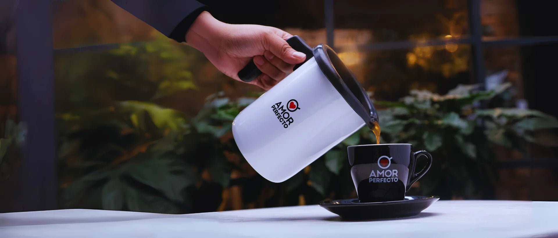 Preparación de café de especialidad Amor Perfecto. Mano sirviendo café recién preparado desde un pichel blanco a una taza negra de la marca, representando los diversos métodos de extracción de café colombiano.